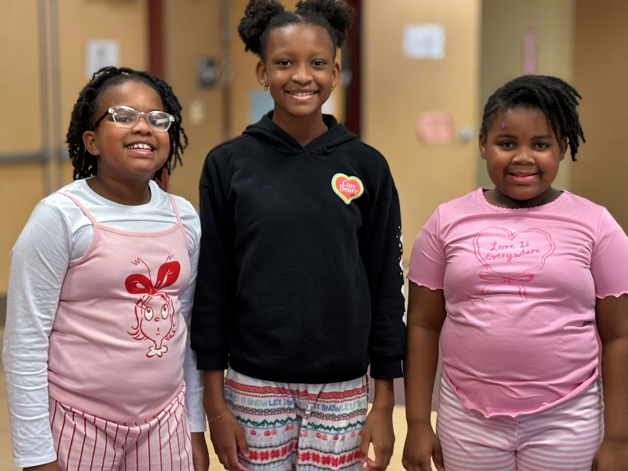 Three smiling girls standing indoors, wearing casual clothes and glasses, with a blurred background.