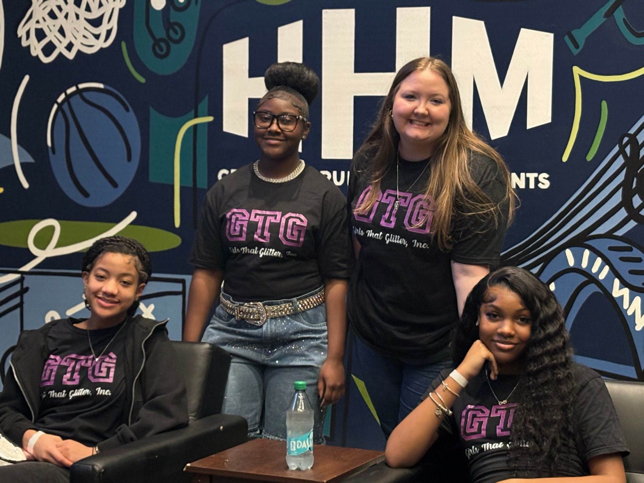Four girls wearing matching black T-shirts with pink text, standing and sitting in front of a colorful sports-themed background.