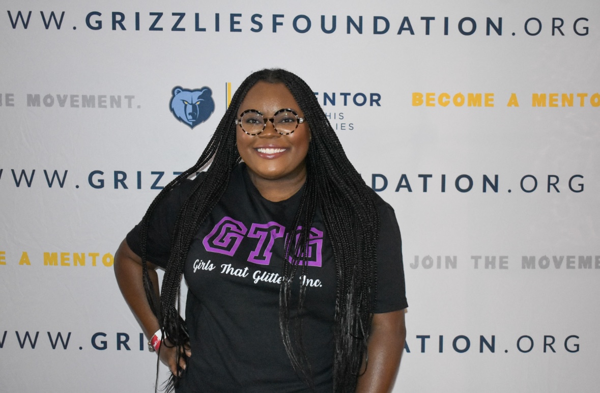 Smiling woman with long braids and glasses standing in front of a backdrop with website and organization logos.