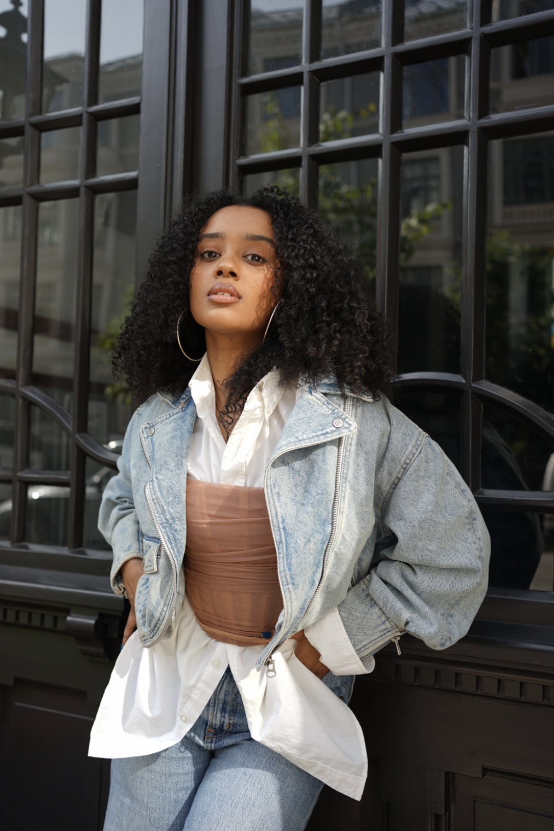Young woman with curly hair wearing a denim jacket and hoop earrings, standing in front of a black grid window.