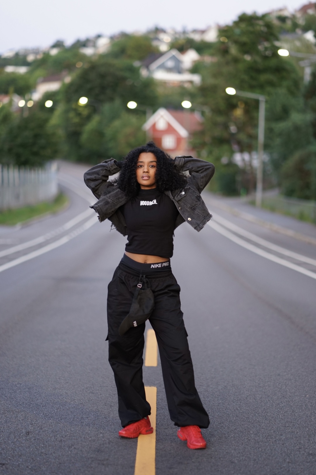 Young woman with curly hair standing in the middle of a road, hands behind head, wearing black outfit and red shoes.