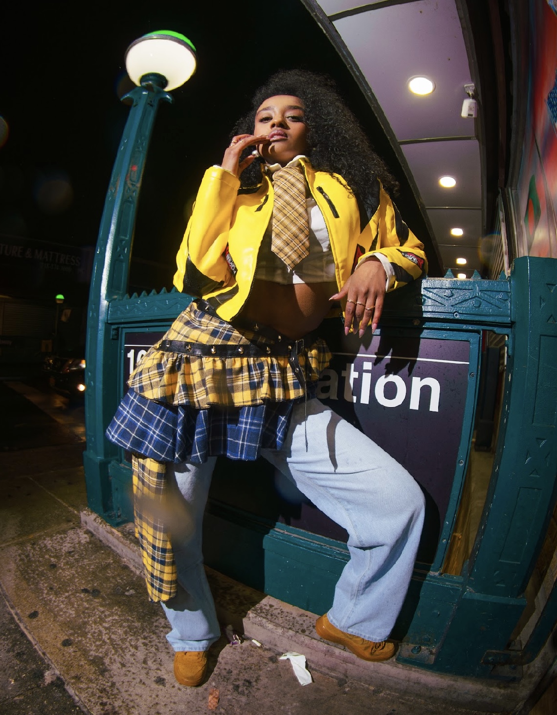 Young woman with curly hair in a yellow jacket, plaid skirt, and light blue jeans, standing outdoors at night.