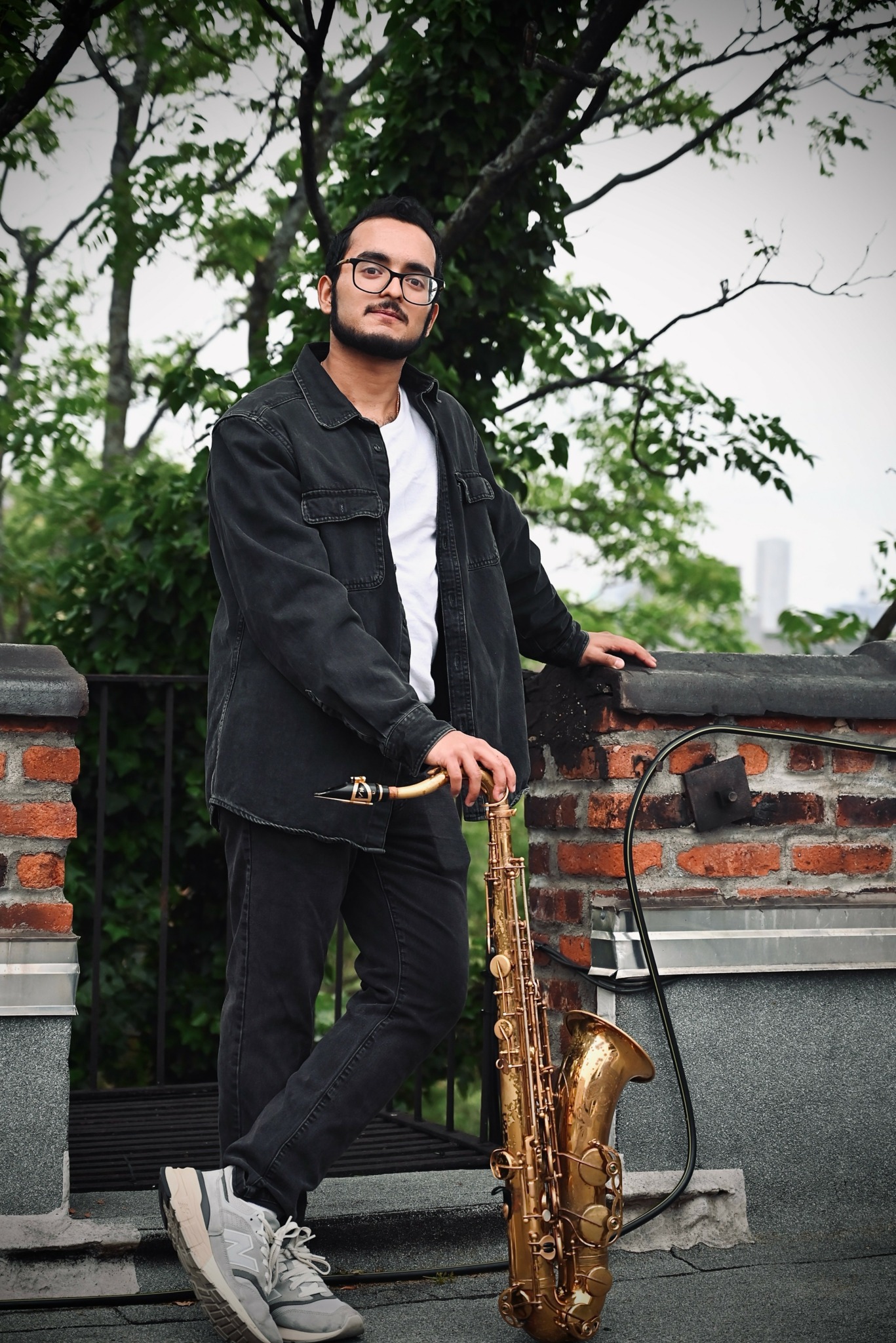 Young man with glasses holding a saxophone outdoors near a brick wall and trees.