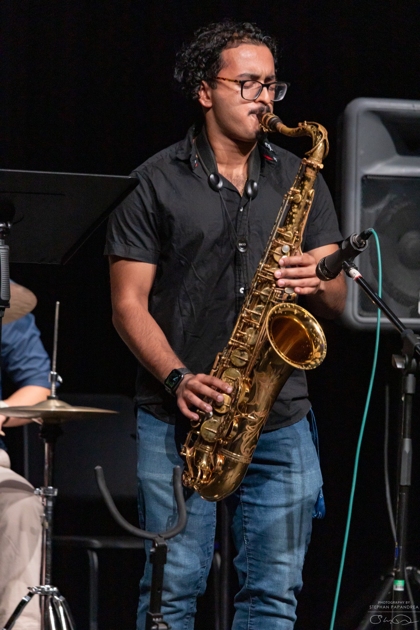 Person playing saxophone on stage with black background, wearing glasses and dark shirt.