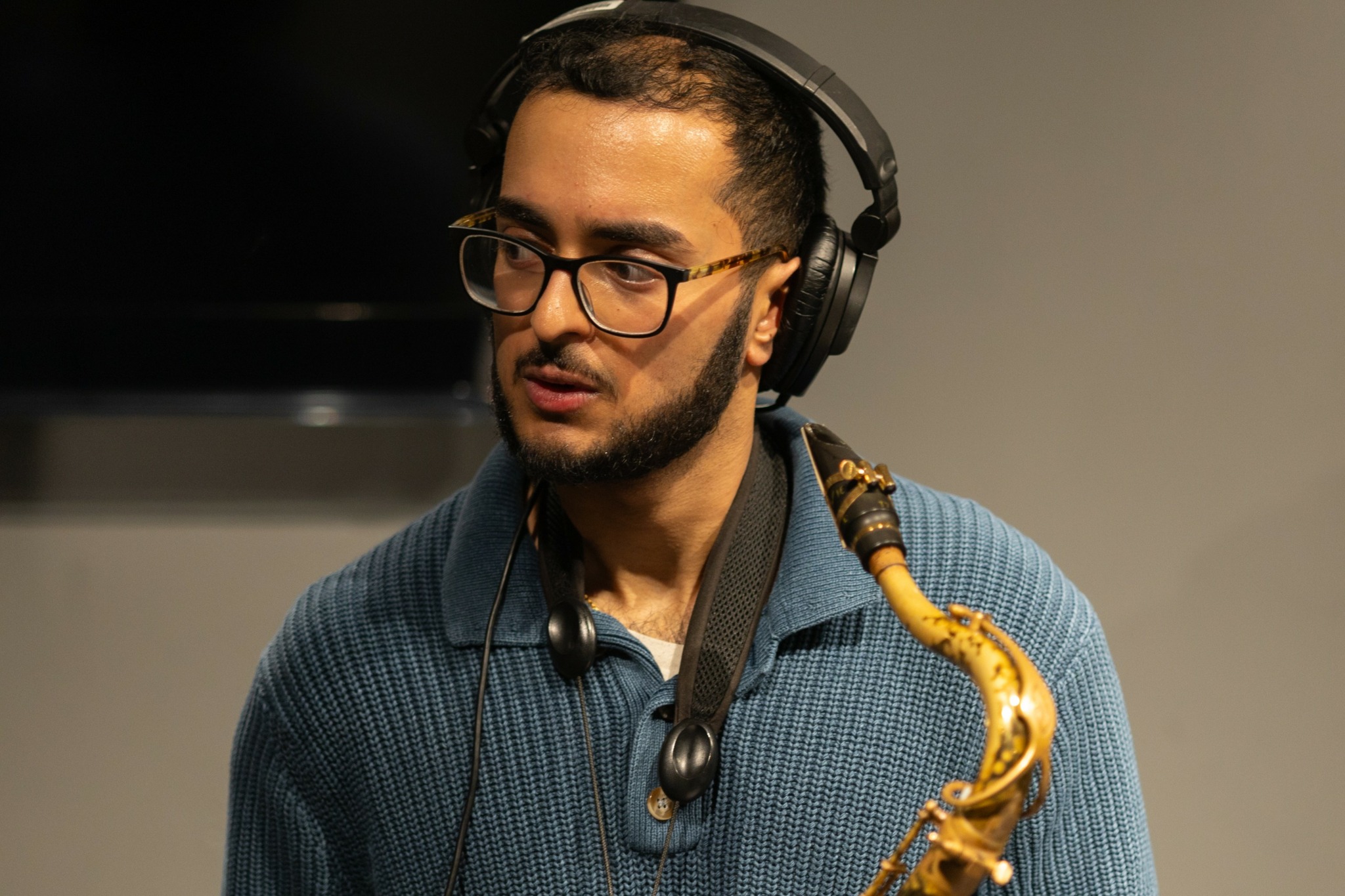 Man with glasses and headphones playing a saxophone, wearing a blue sweater, in an indoor setting.