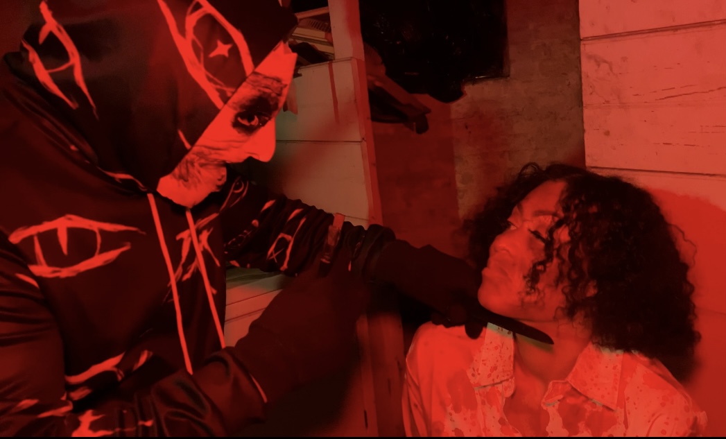 Person with curly hair sitting and another person leaning towards them in a red-lit environment.