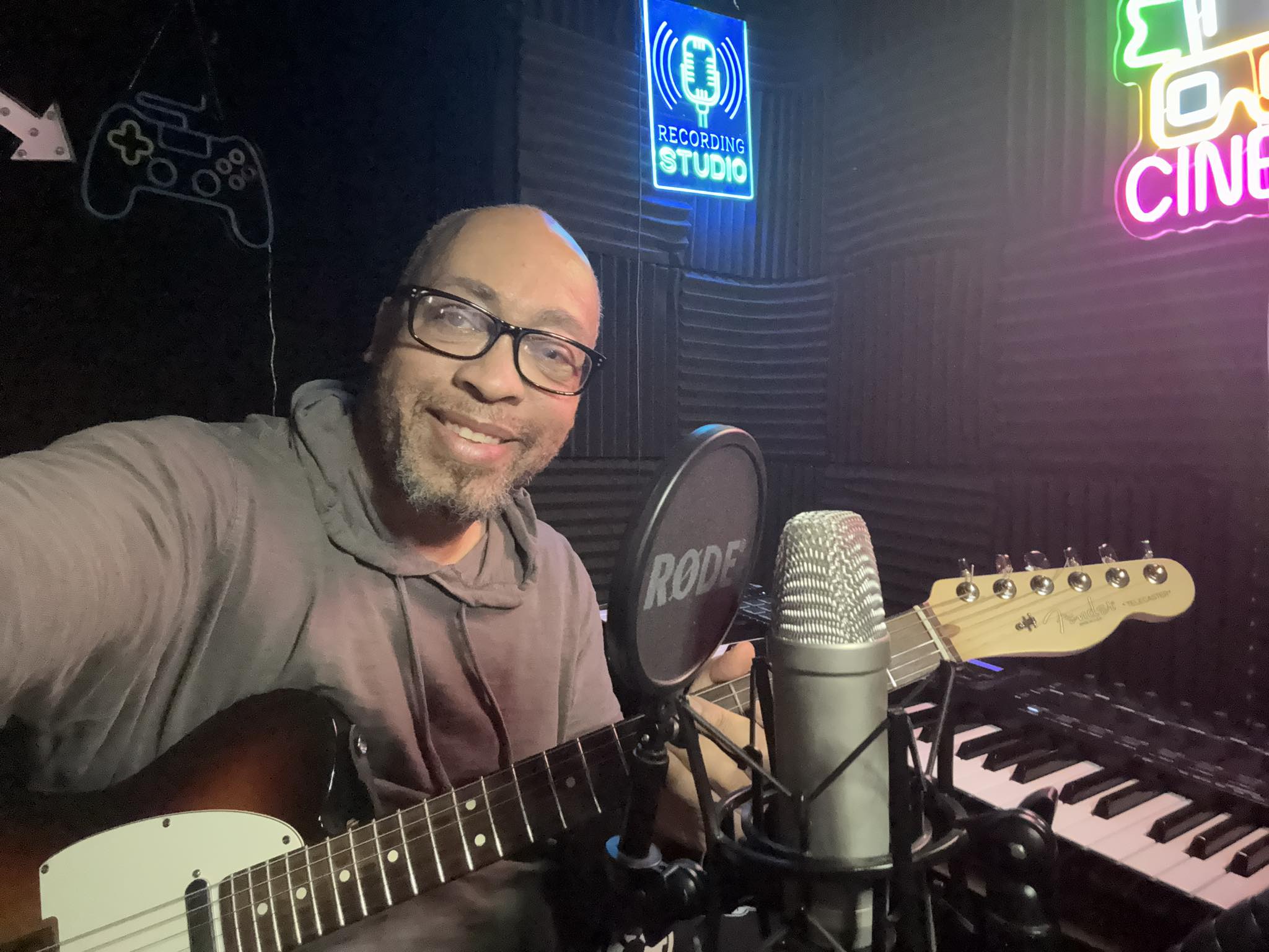 Person with glasses smiling, holding guitar in recording studio with neon signs and keyboard in background.