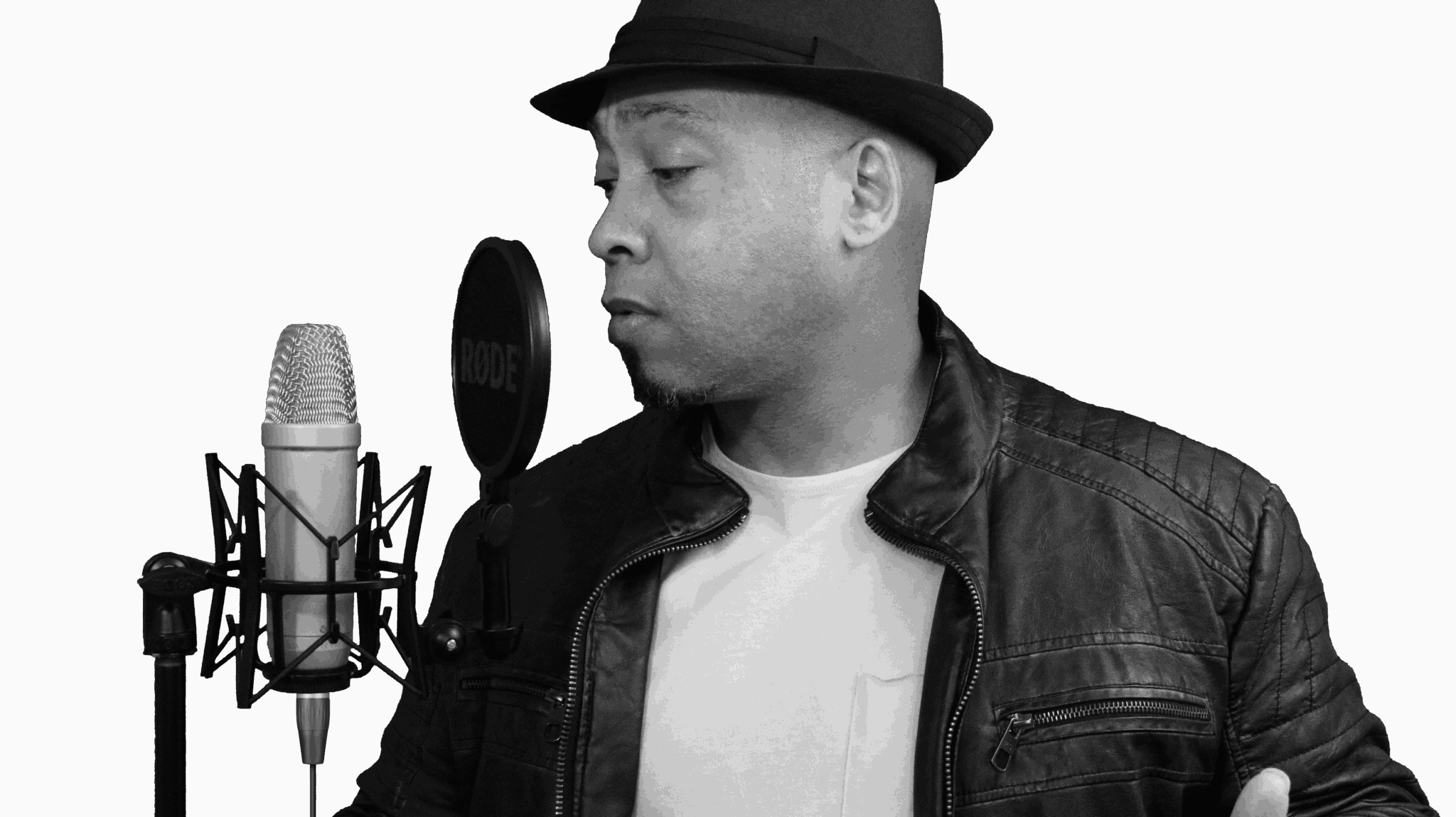 Man wearing a hat and leather jacket singing into a microphone in a dark studio.