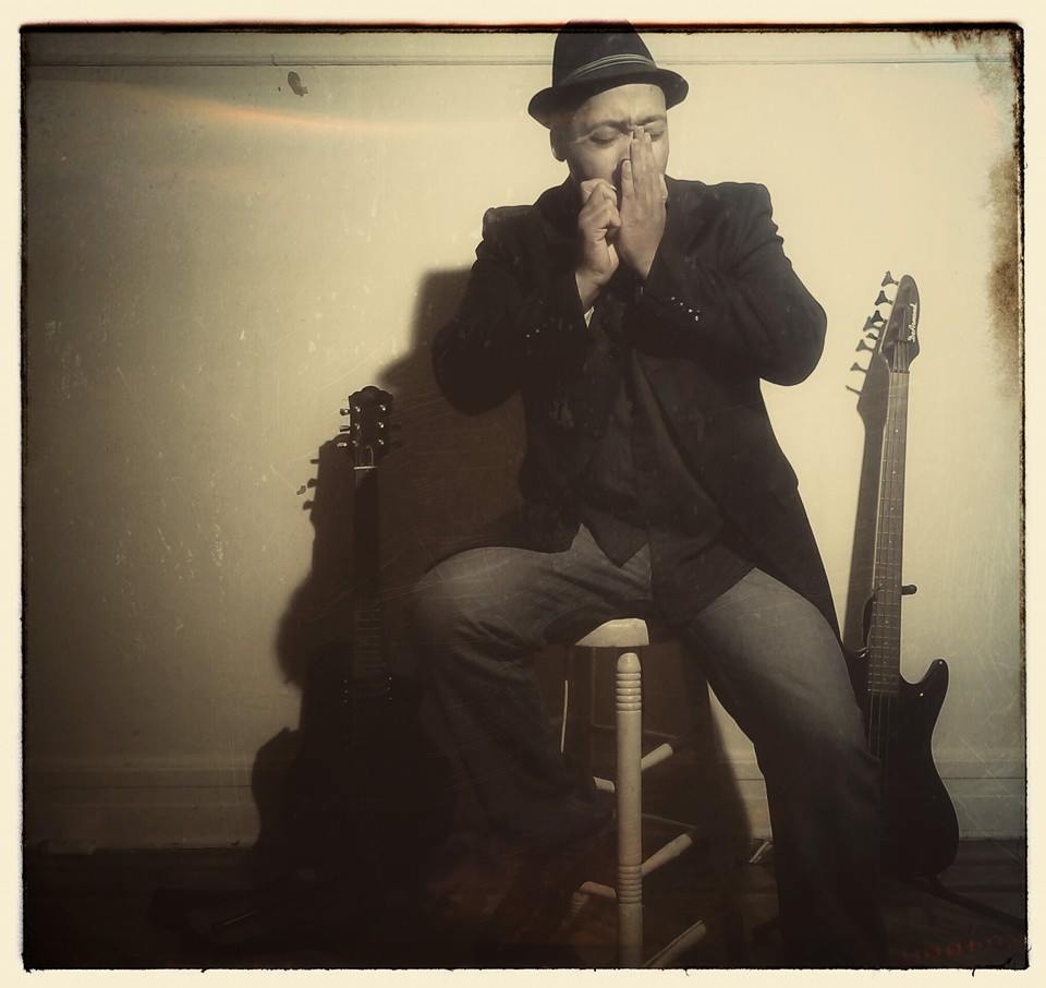 Person sitting on a stool with hands covering mouth, wearing a hat and glasses, two guitars beside them.