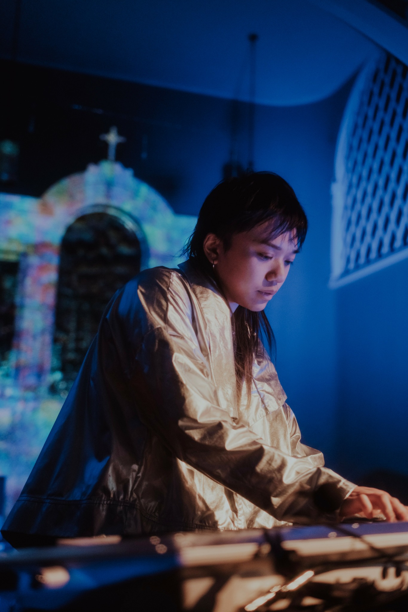 Young woman with long dark hair wearing a shiny gold jacket, focused on a keyboard or musical instrument.