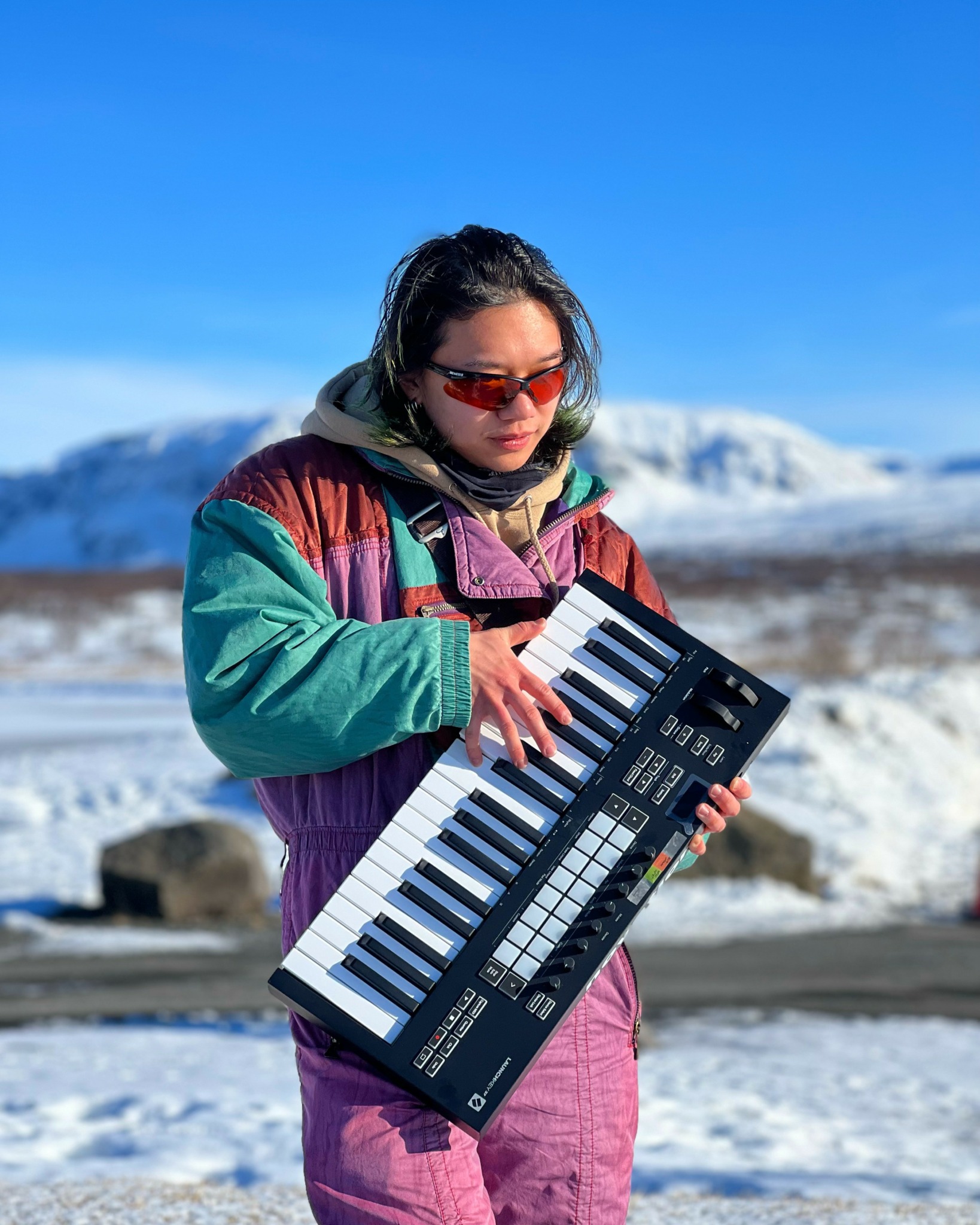 Person wearing sunglasses and colorful jacket playing a keyboard outdoors with snowy mountains in background.