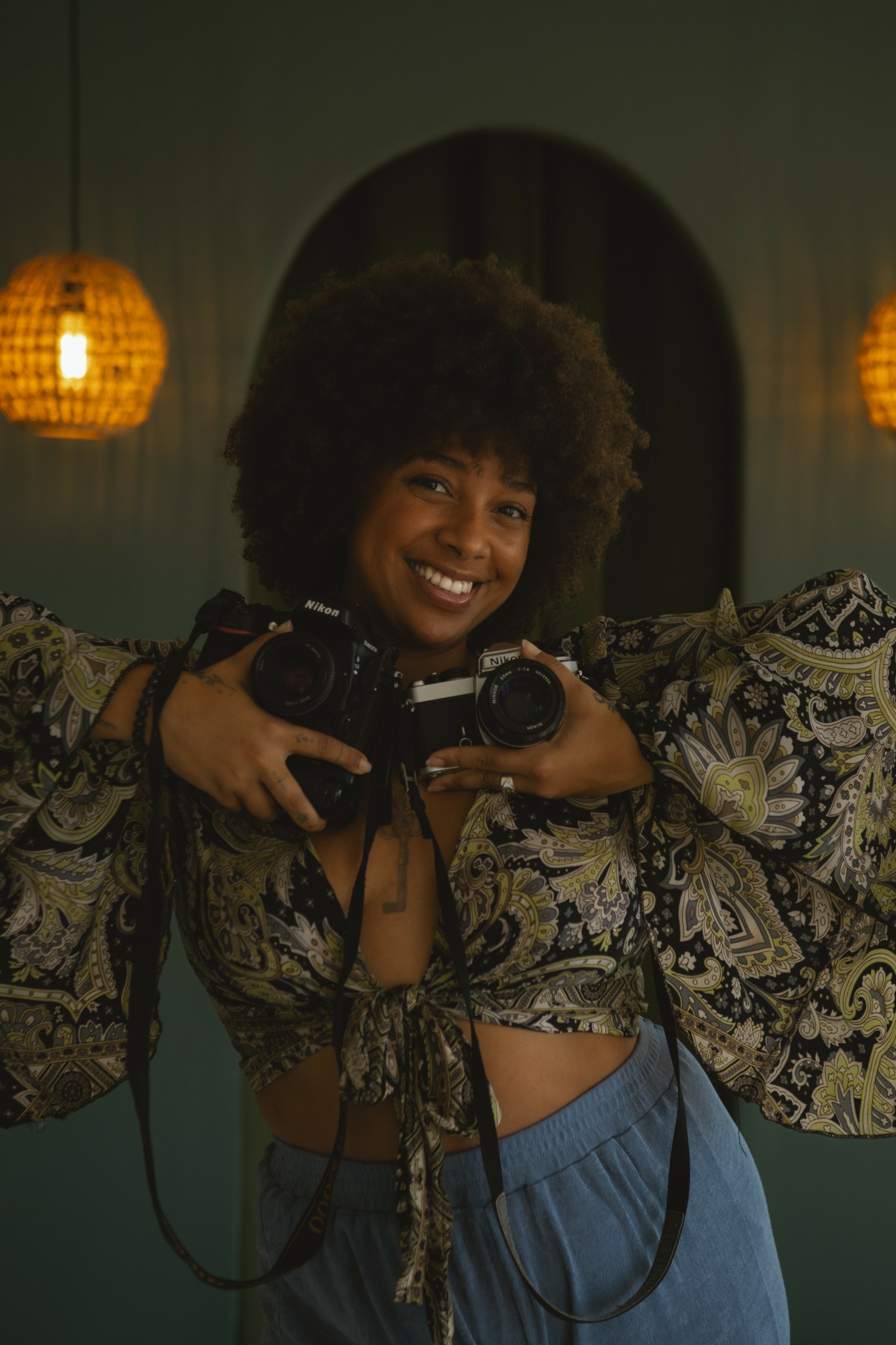 Person with curly hair smiling, holding a camera, wearing patterned clothing, in a dimly lit room with hanging lamps.