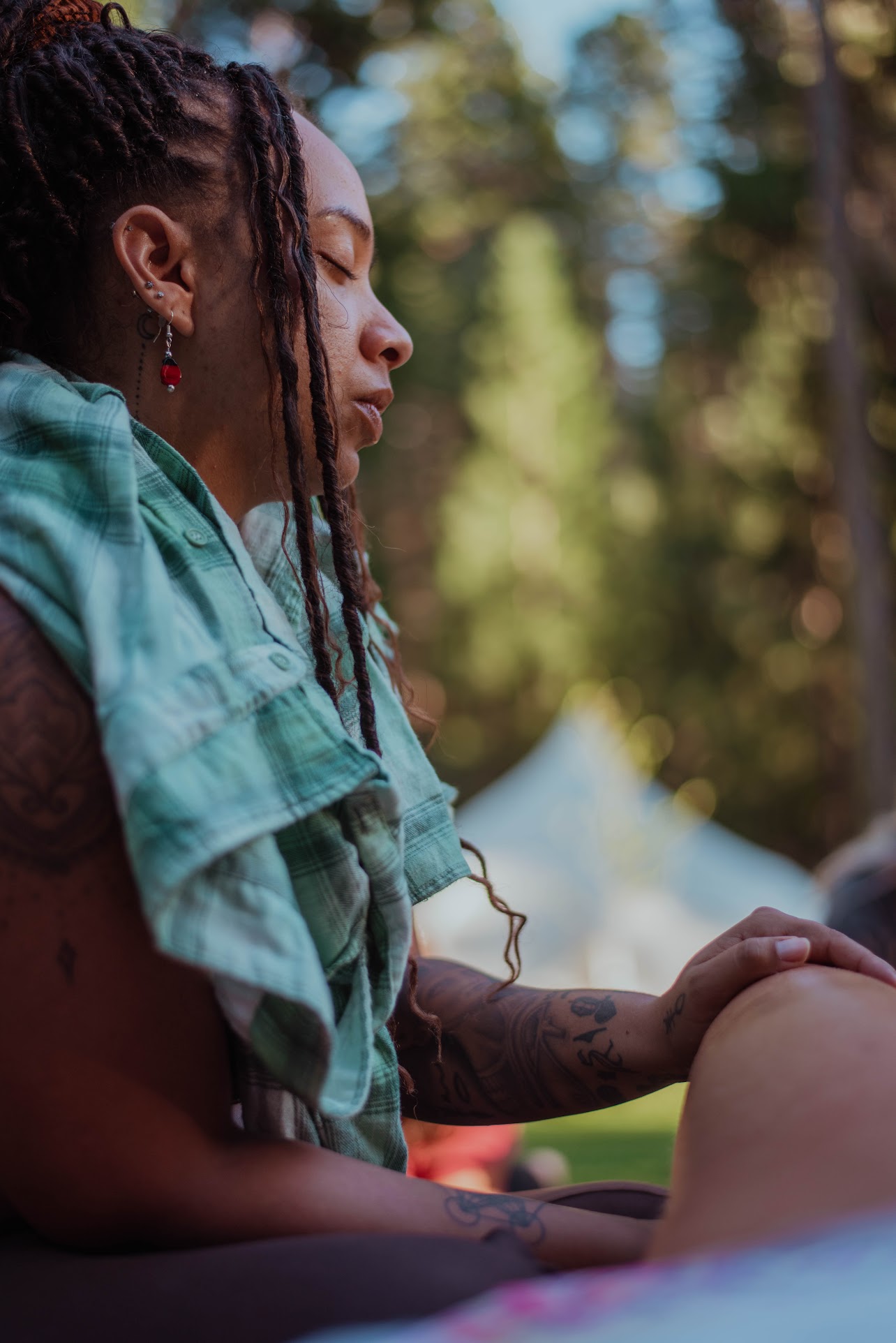 Side profile of a person with dreadlocks outdoors, with trees and sunlight in the background.