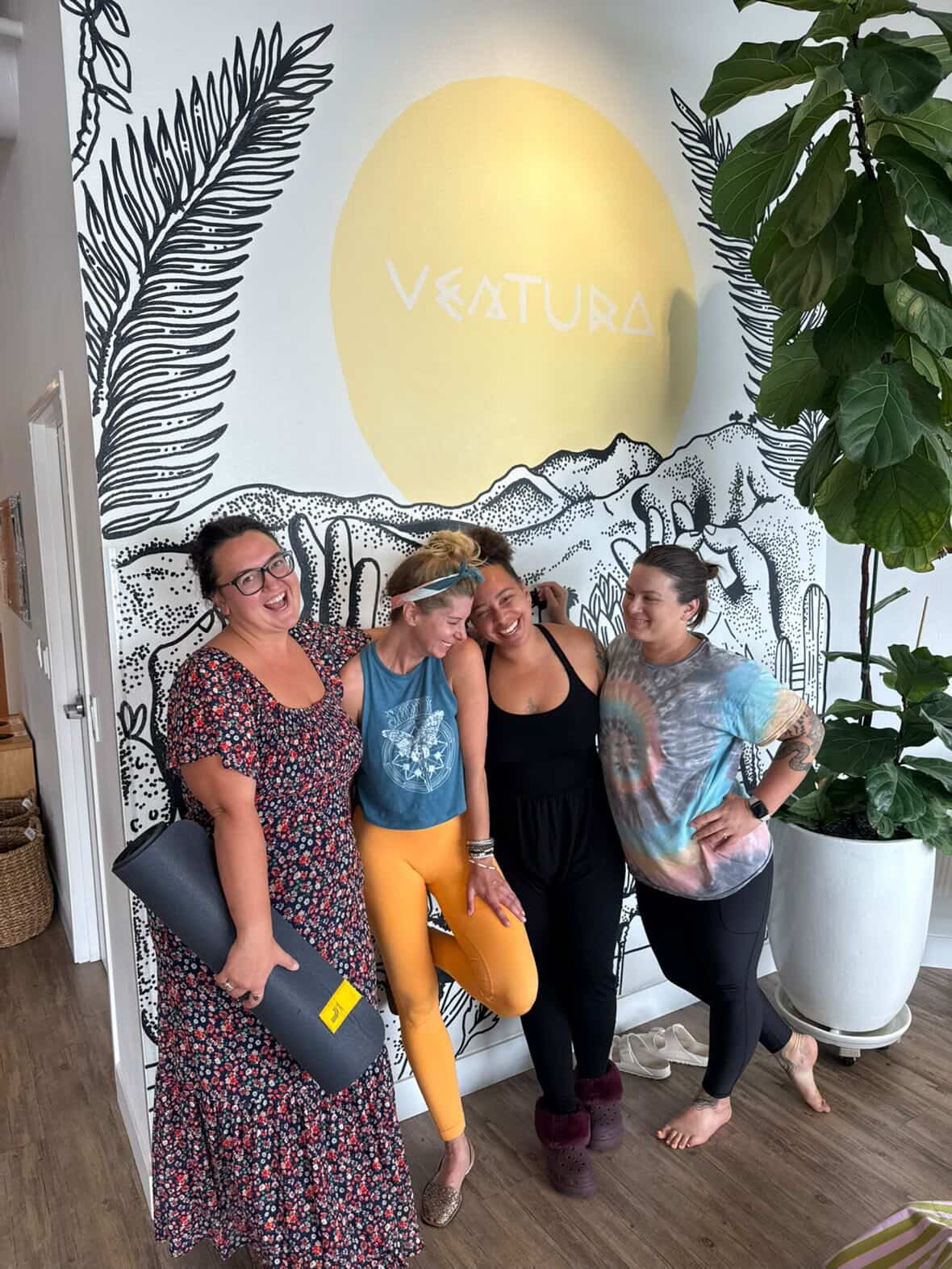 Four women standing together in front of a mural with a yellow circle and mountain scene, smiling and posing.