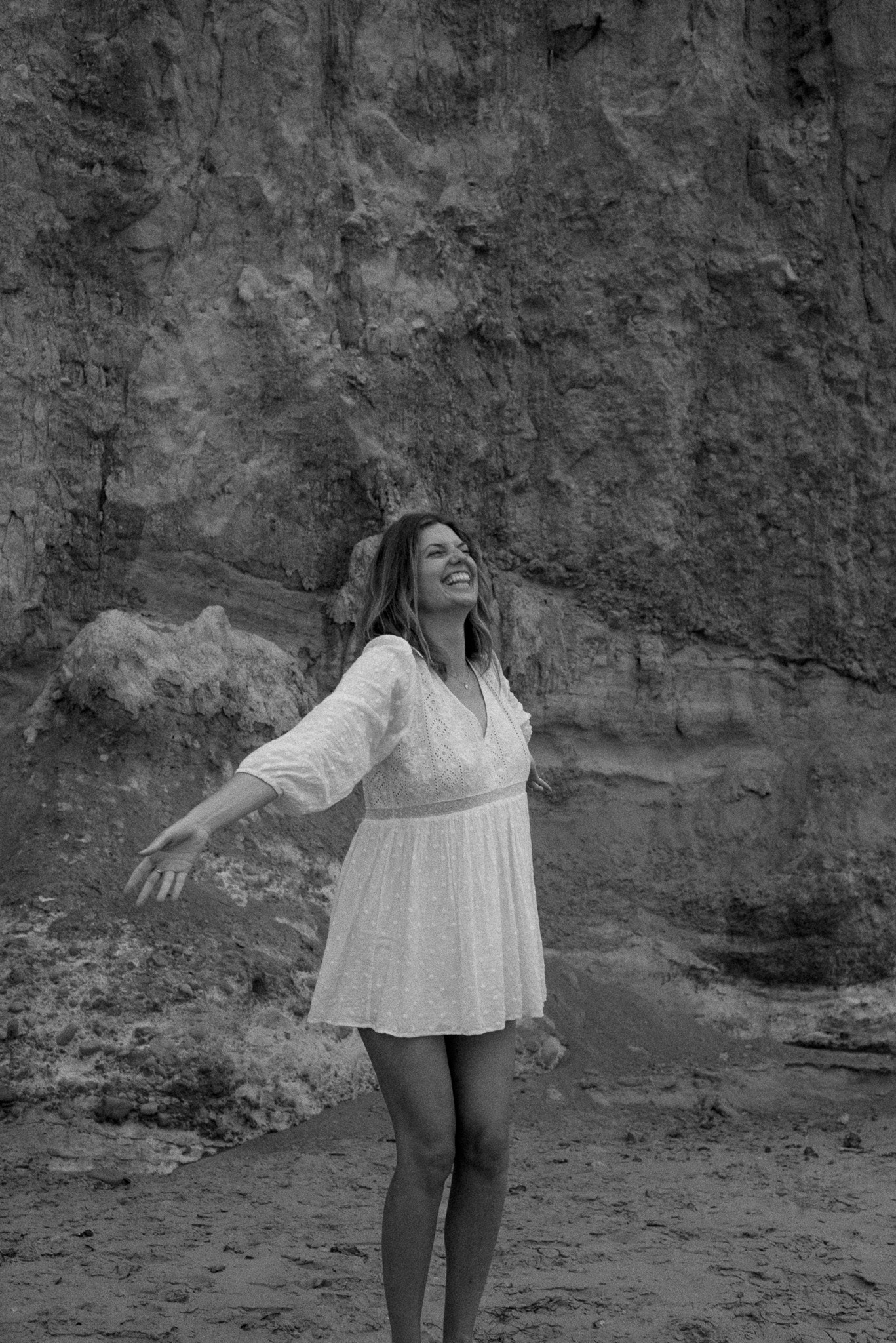 Woman standing outdoors with arms outstretched, smiling, in front of a rocky cliff.