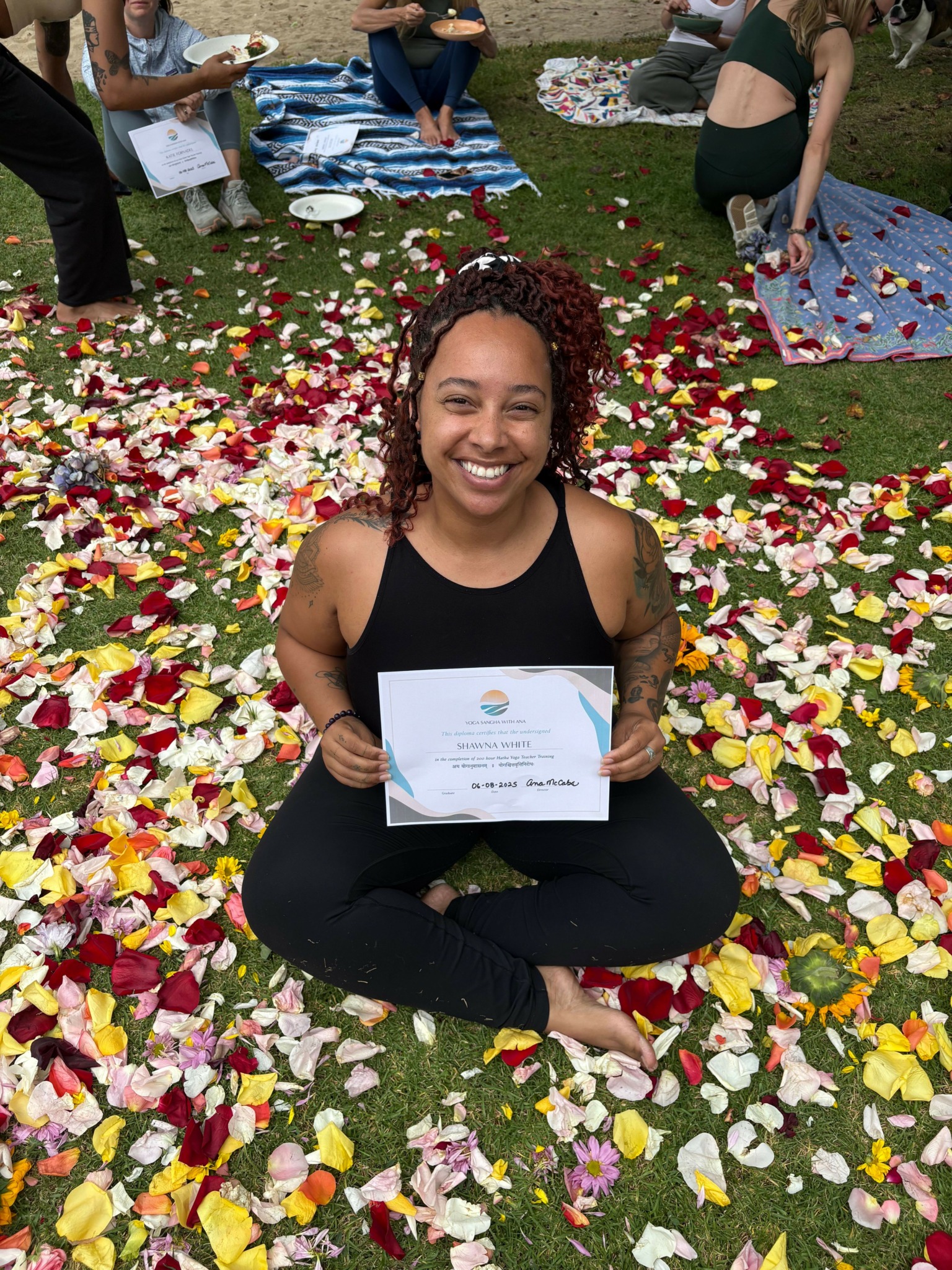 Woman sitting on flower petals holding a certificate, surrounded by colorful flower petals on grass.