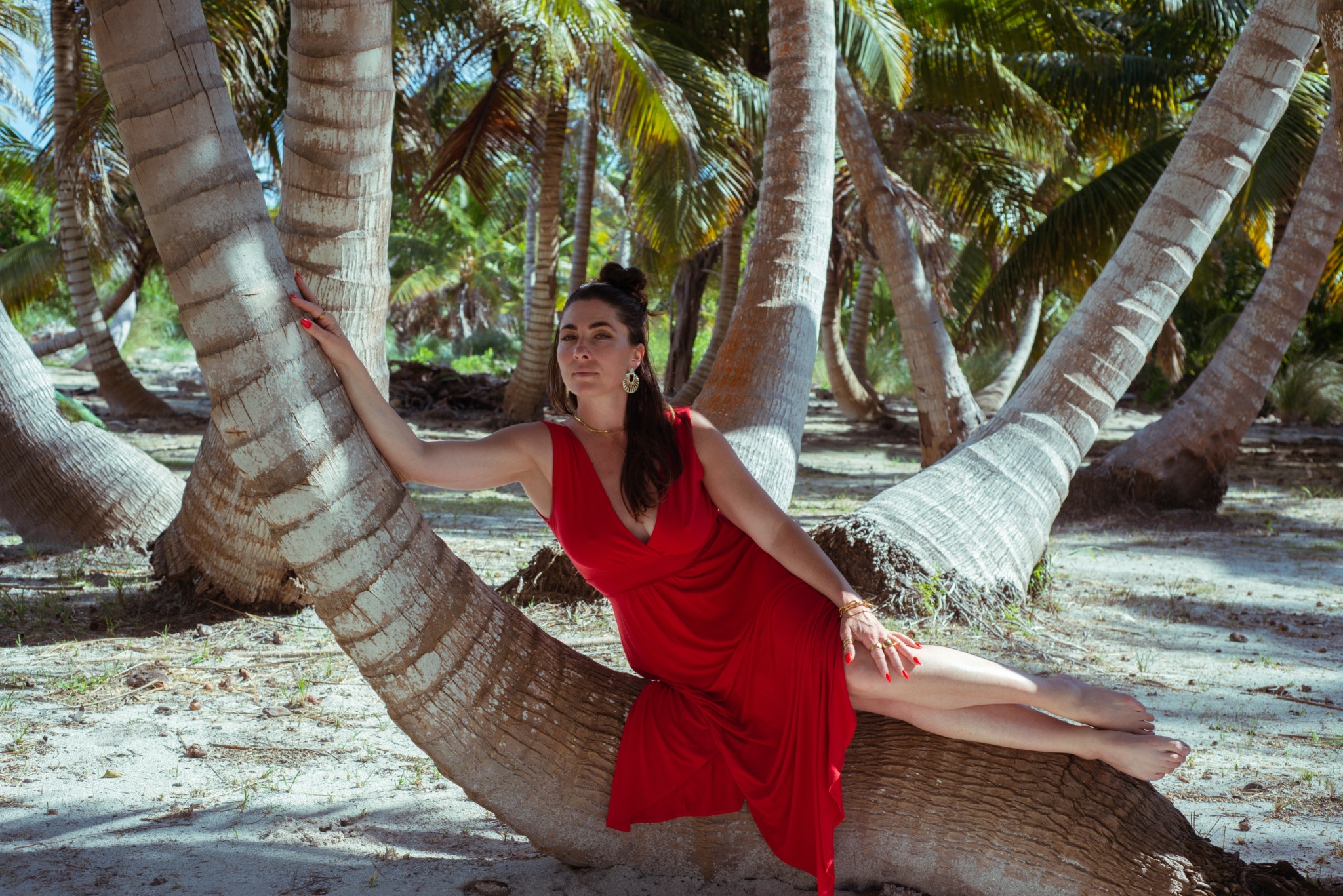 Woman in red dress lying on a tree trunk among palm trees on a sandy beach.