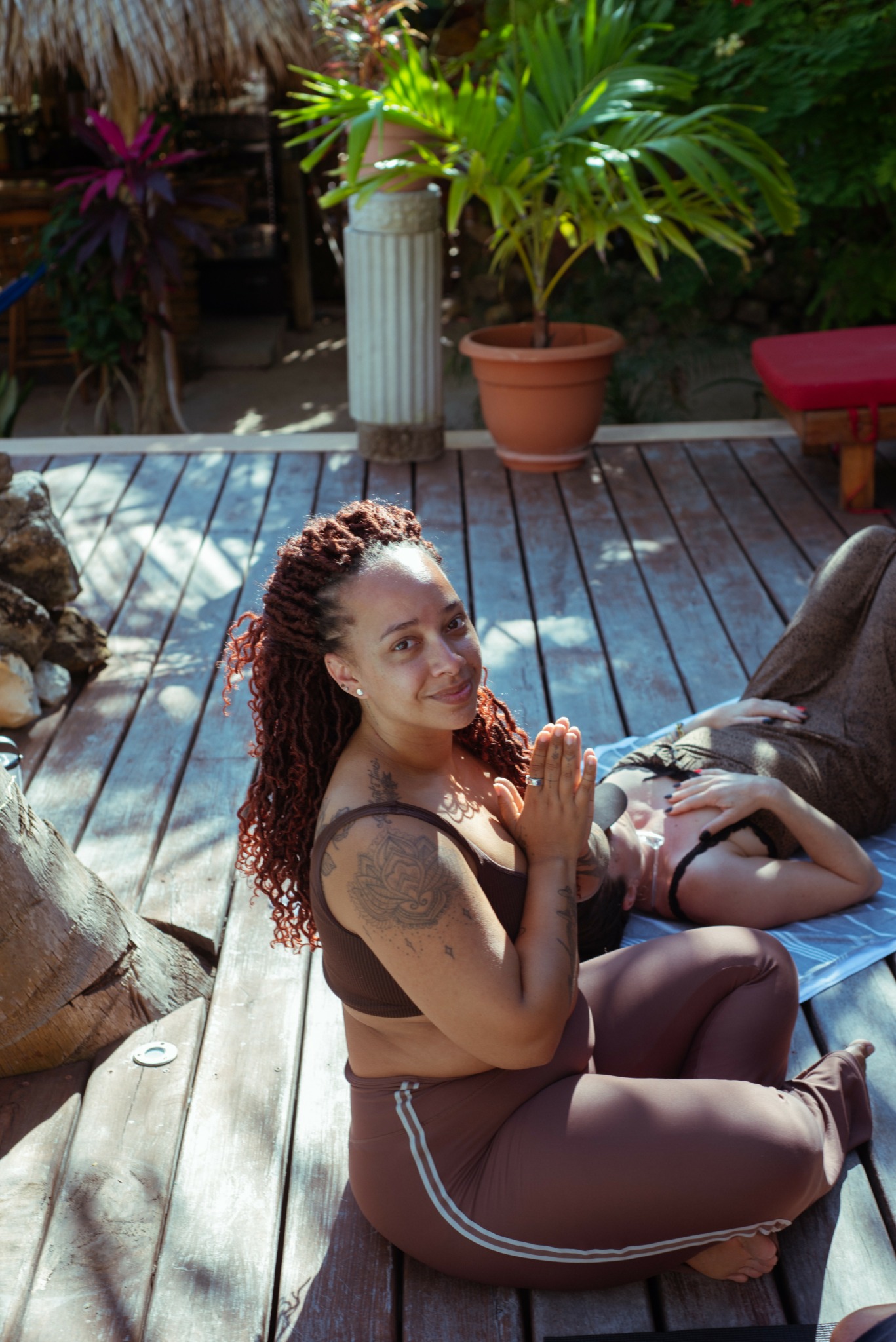 Woman with curly hair sitting on wooden deck, smiling, with potted plants and a person lying nearby.