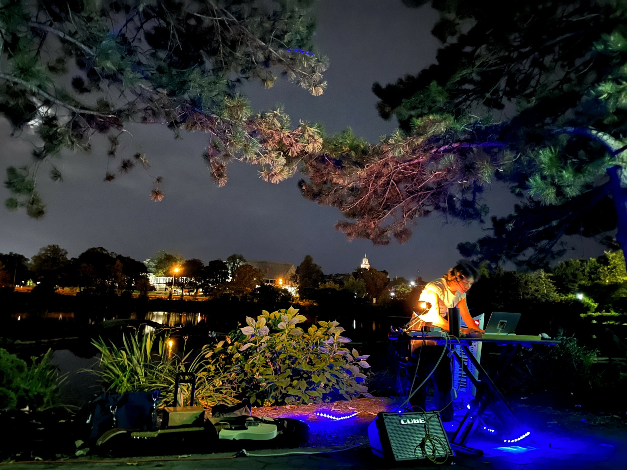 Person performing music outdoors at night near a body of water with trees and lights reflected in the water.