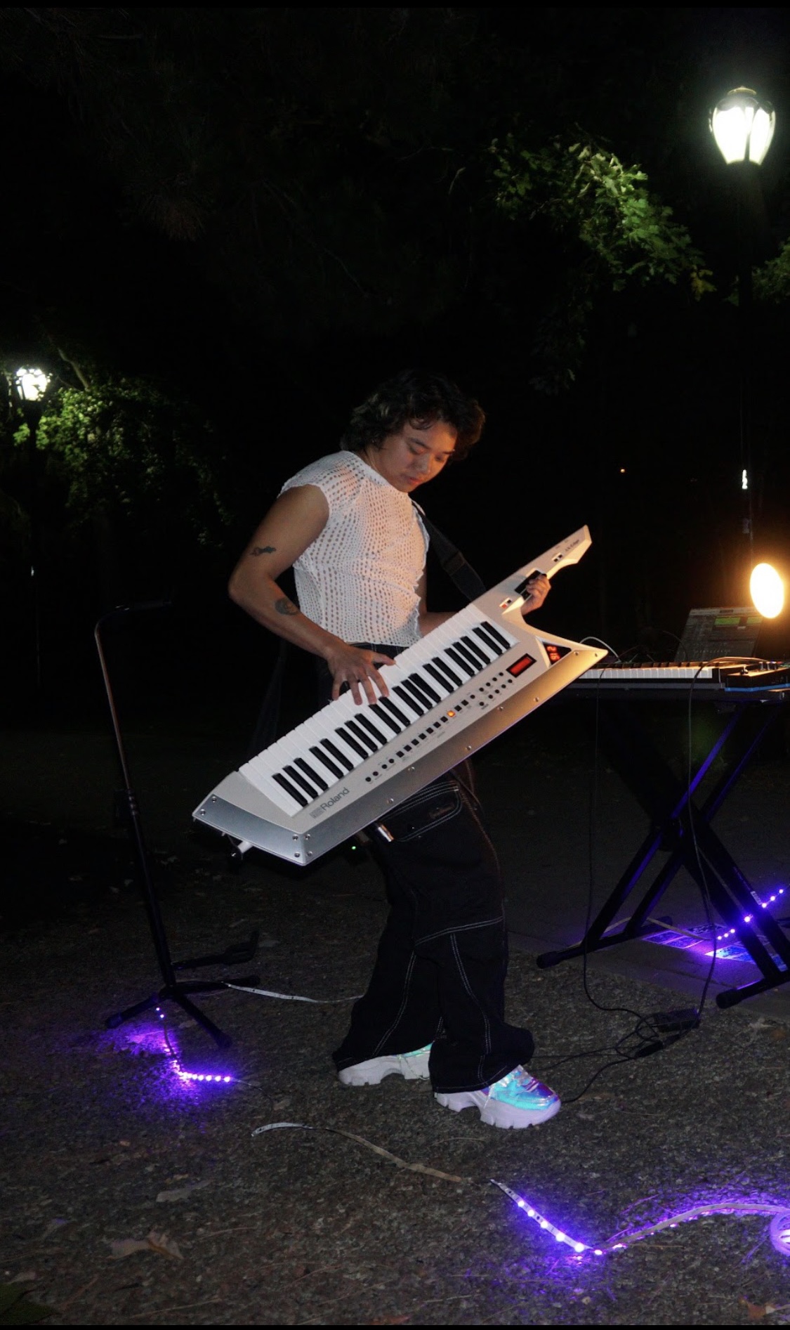 Person playing a keyboard outdoors at night, illuminated by streetlights, with purple lights on the ground.