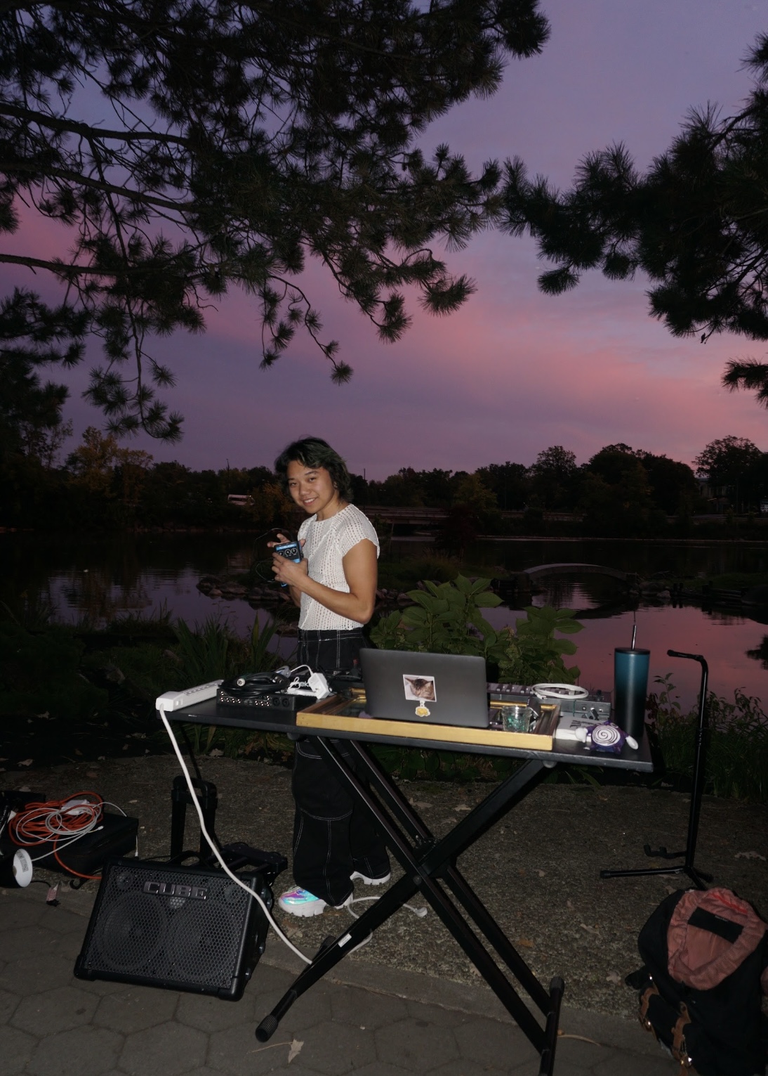 Person standing outdoors at sunset with a laptop and music equipment on a table, near a body of water and trees.