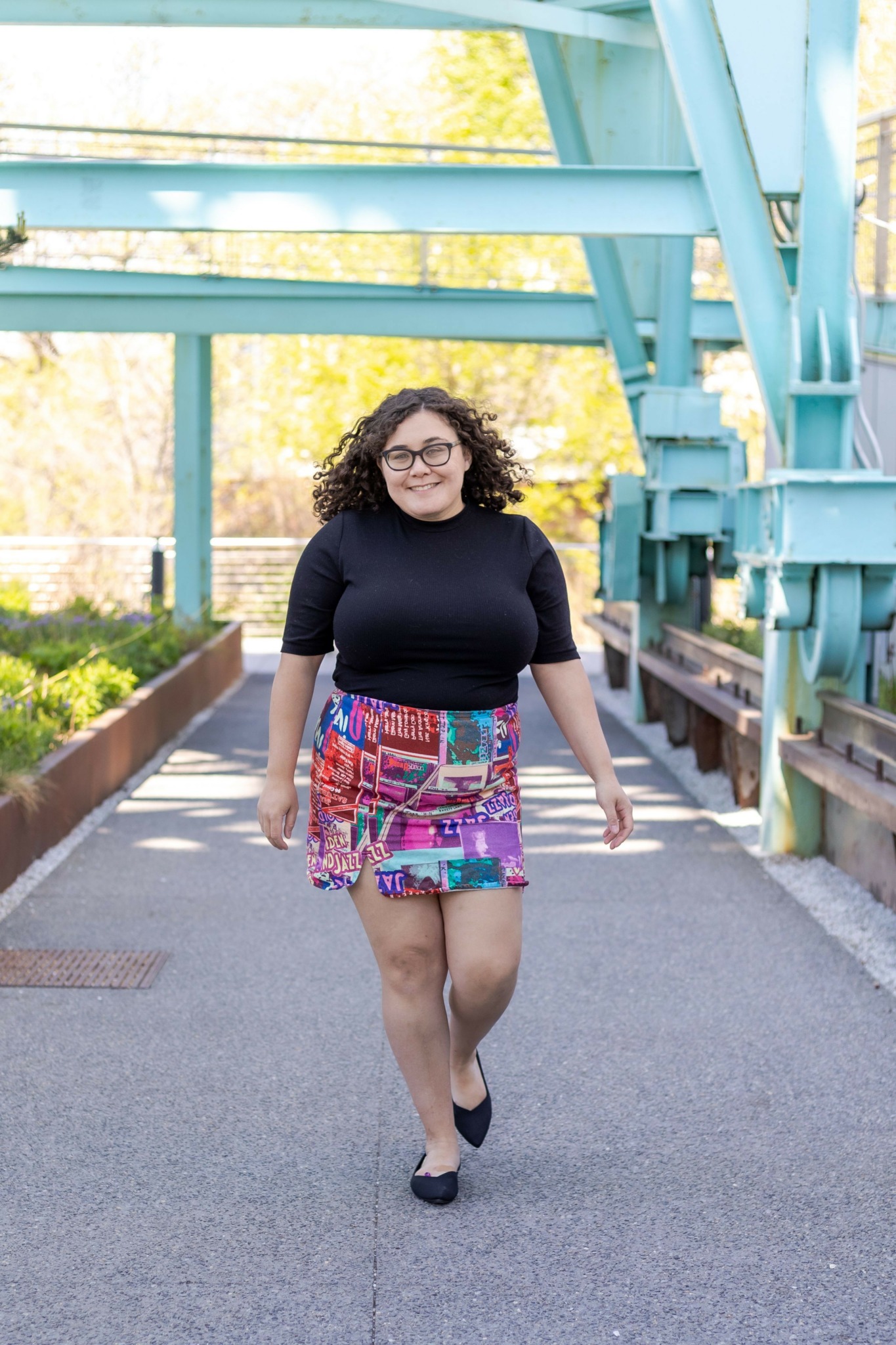 Woman standing outdoors on a paved path near a blue structure, wearing a black top and colorful skirt.