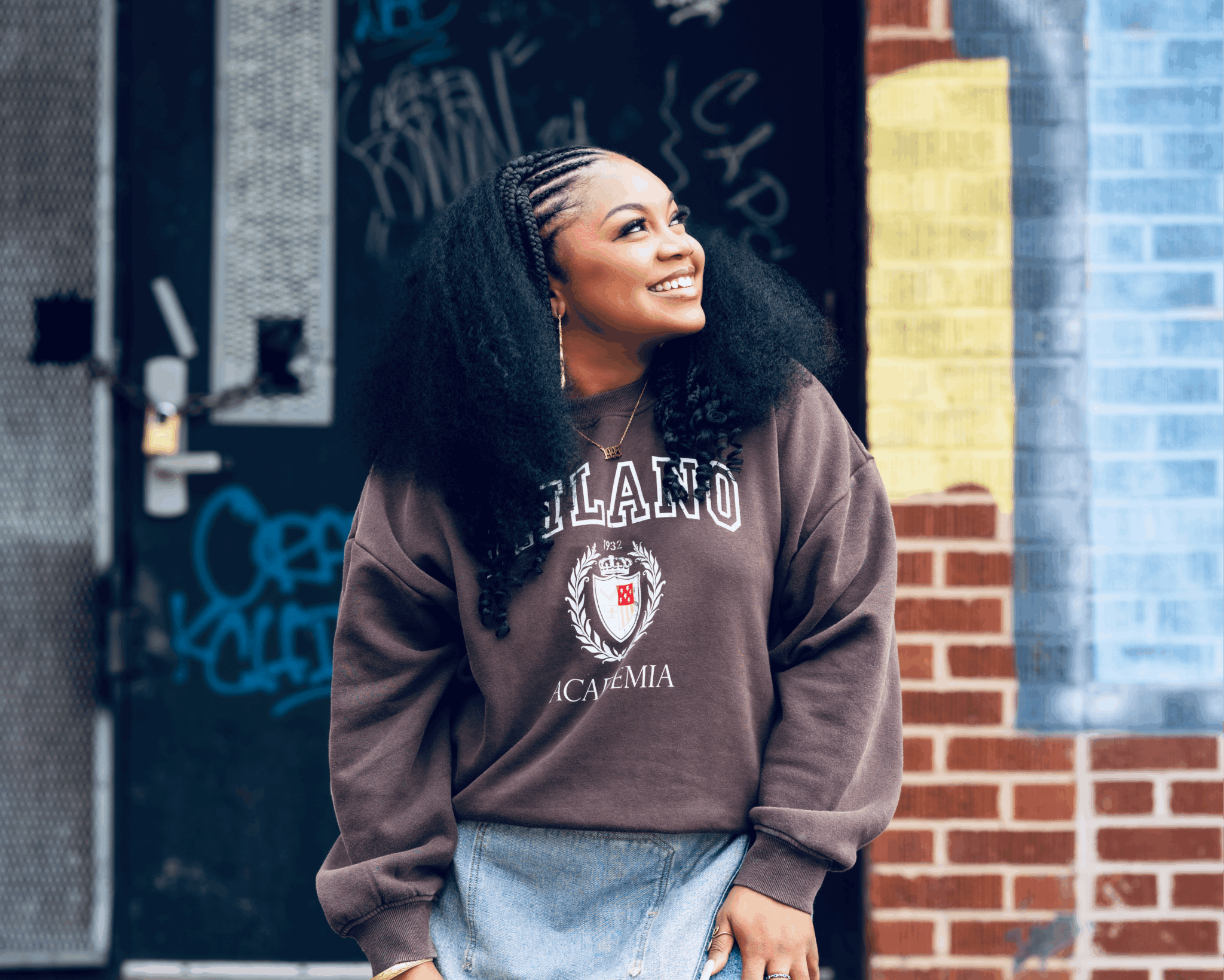 Smiling woman with curly hair stands outdoors near a brick wall and graffiti.