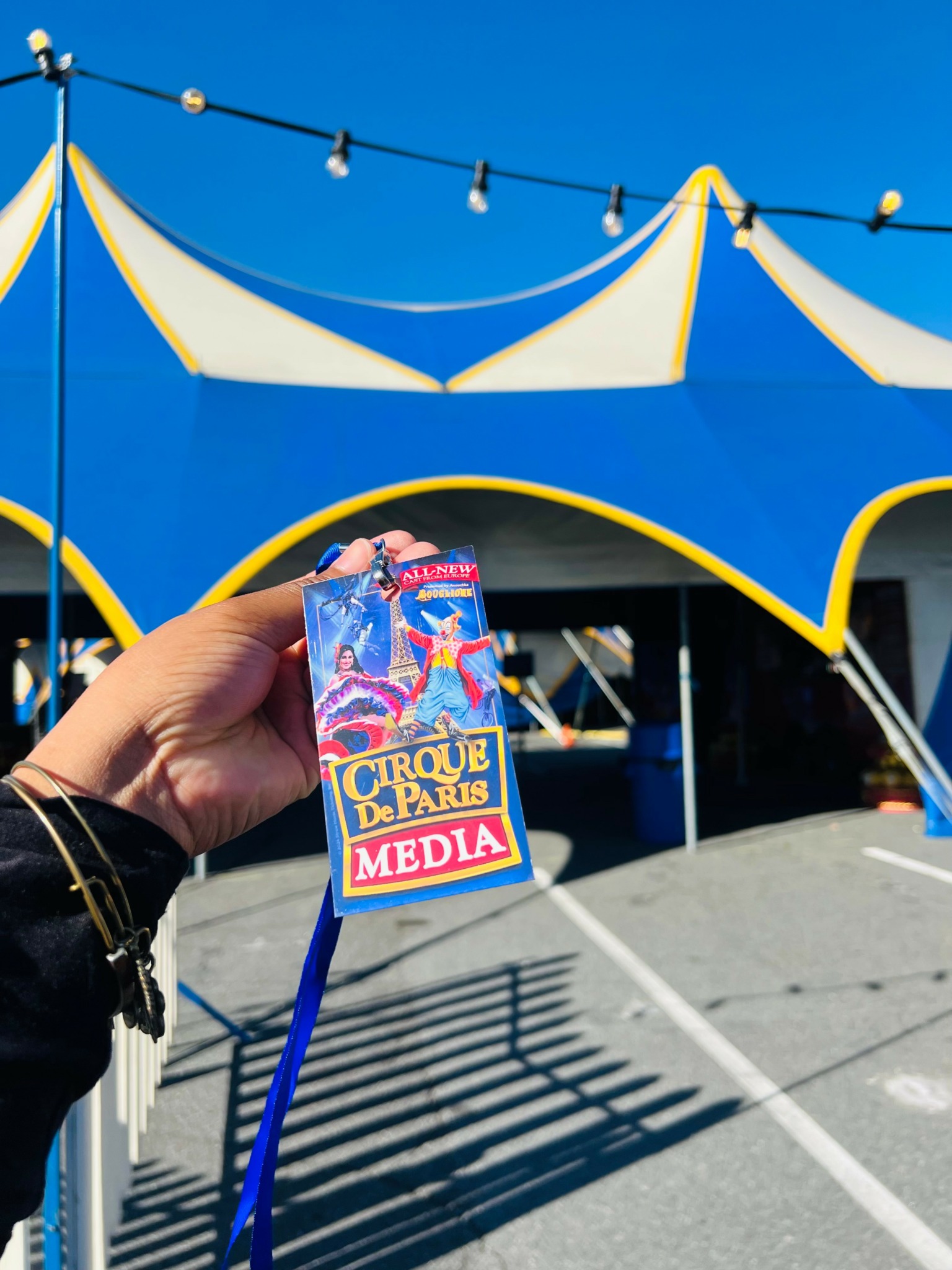 Person holding a colorful circus ticket in front of a blue and yellow circus tent under a clear sky.