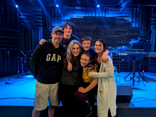 Group of six people smiling on stage with musical instruments and equipment in background.