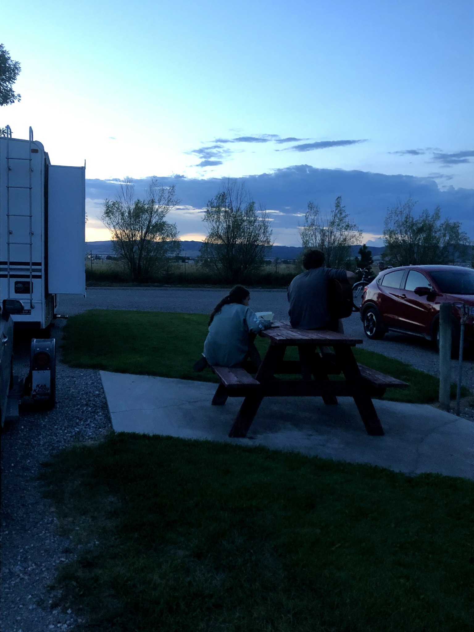 Two people sitting at a picnic table outdoors during dusk, with a truck and parked car nearby, trees in the background.