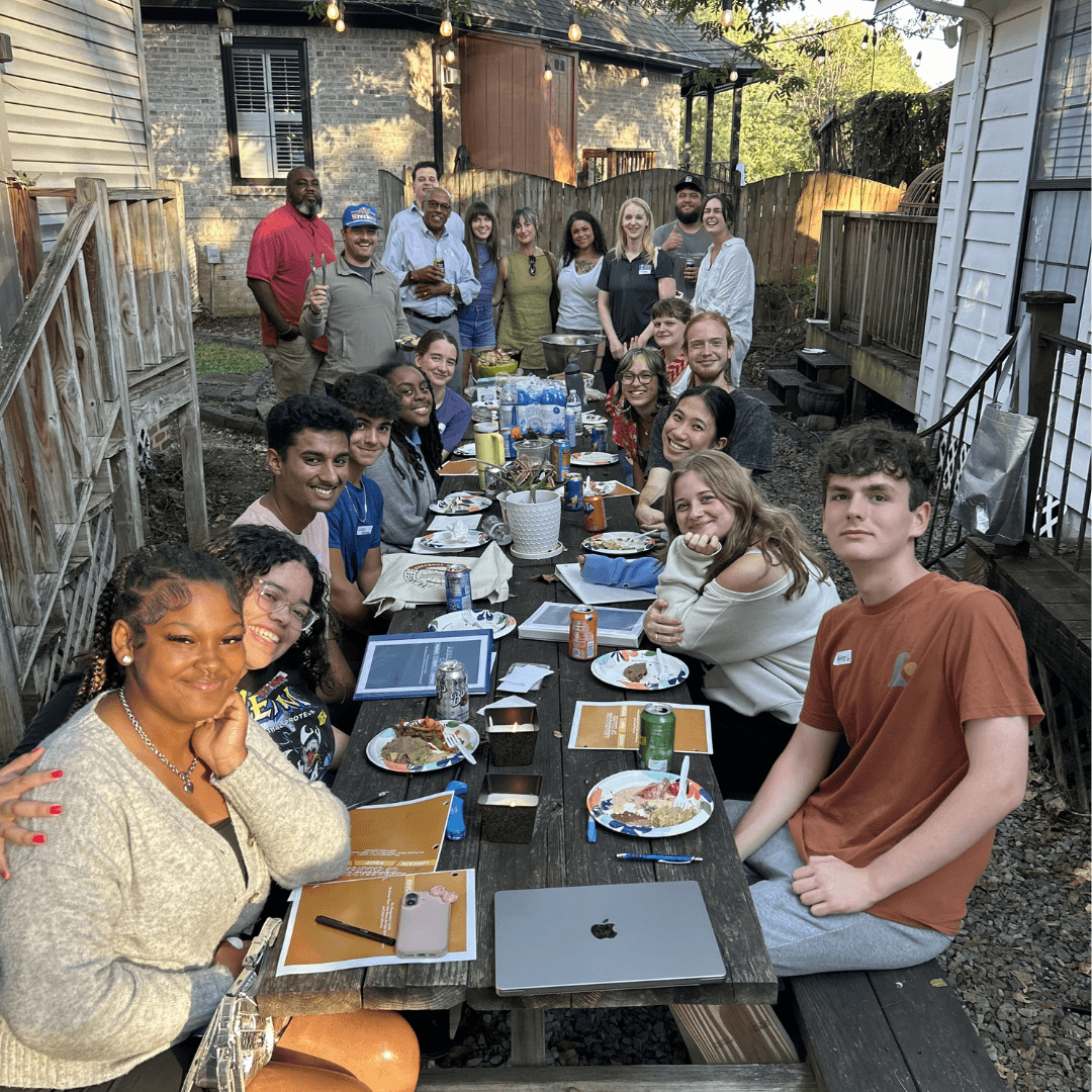 Group of people gathered around a long outdoor table with food and drinks, smiling and posing for the photo.