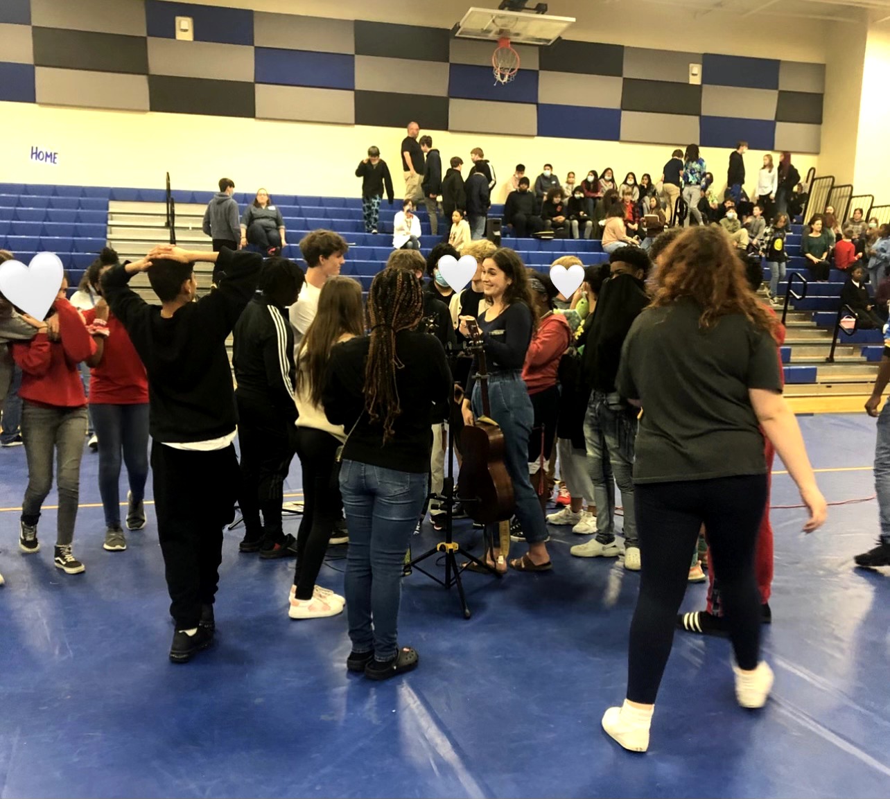 Group of people gathered in a gymnasium, some holding musical instruments, with bleachers and a basketball hoop in background.