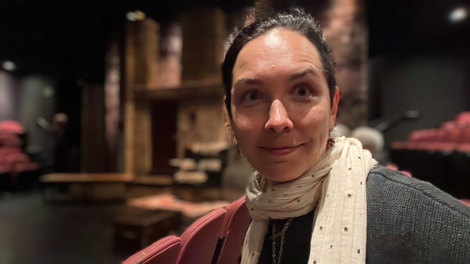 Woman with dark hair tied back, wearing a scarf and dark top, in a dimly lit indoor setting.