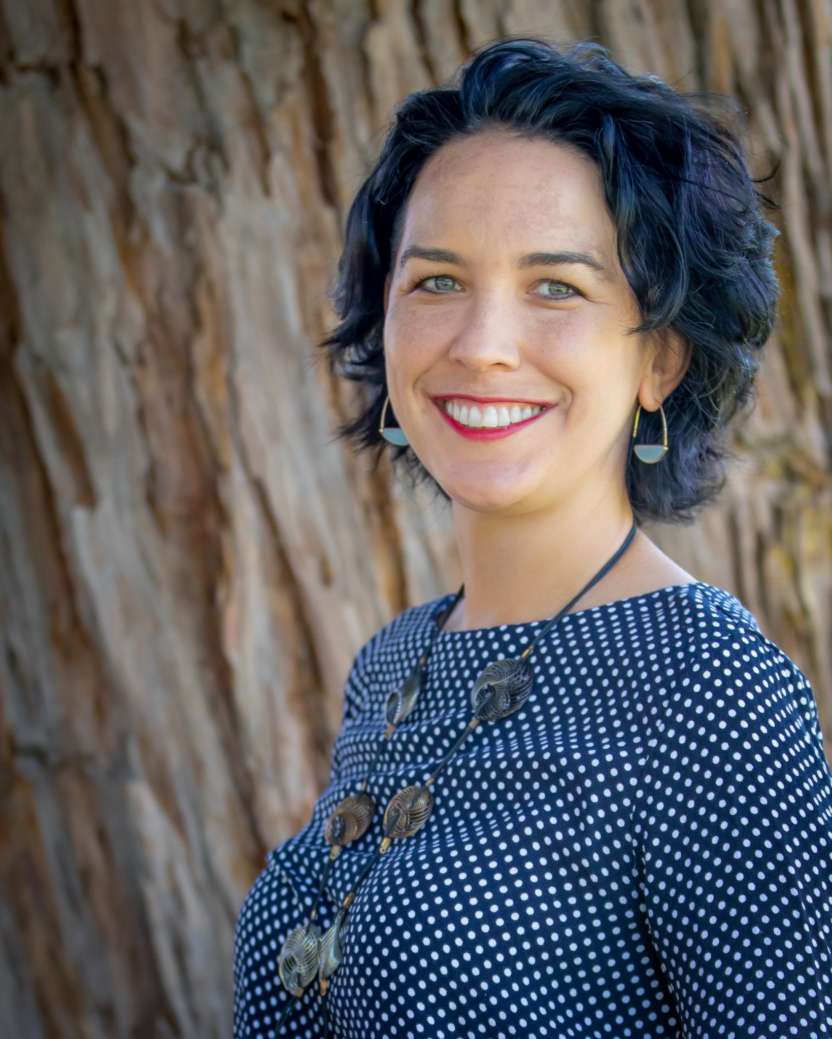 Woman with short dark blue hair smiling outdoors, wearing a polka dot top and earrings.