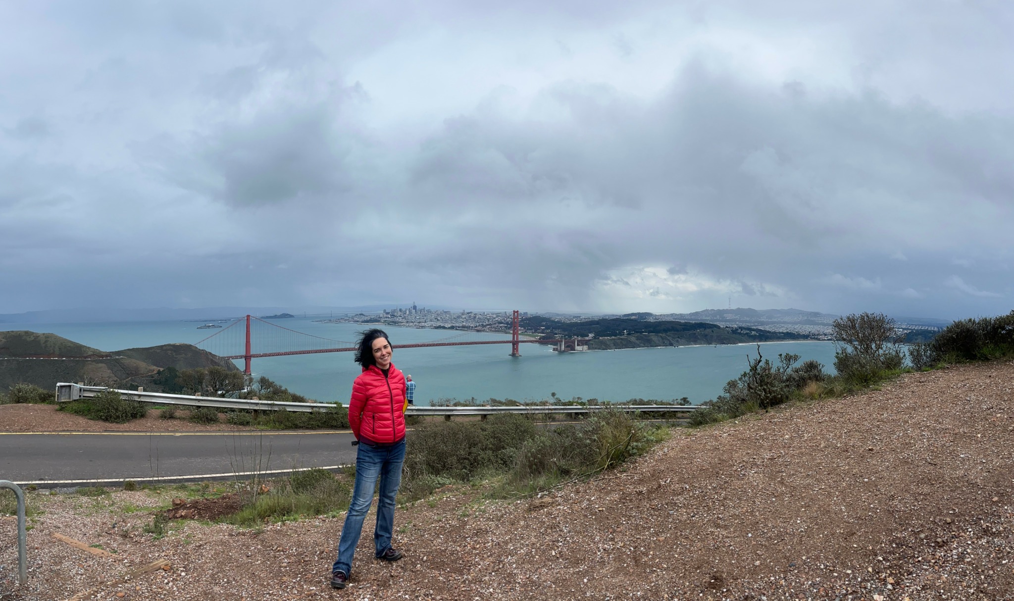 Person standing outdoors near a road with a bridge and water in the background under cloudy sky.