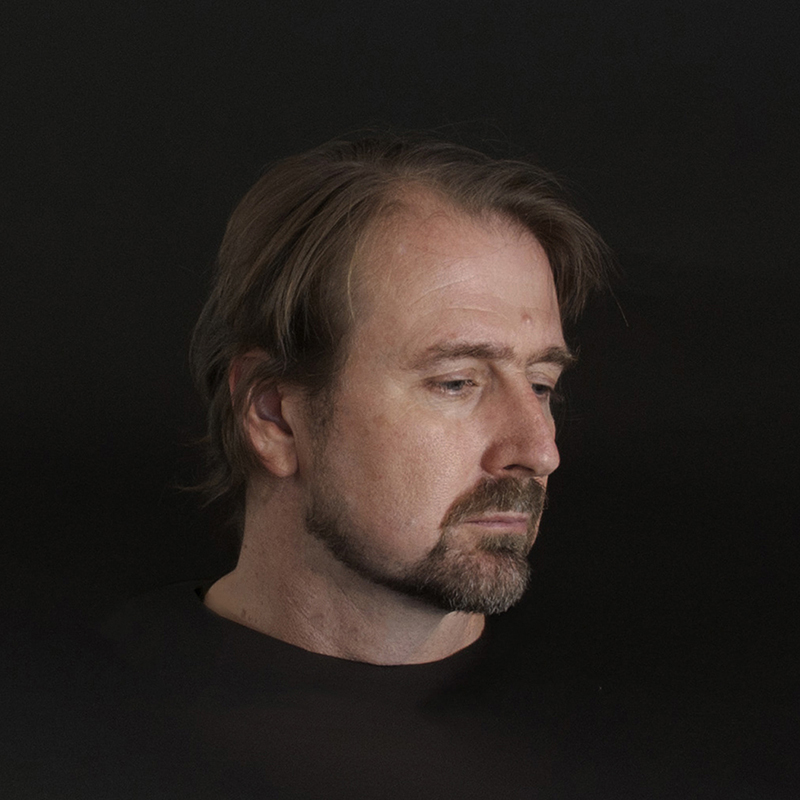 Side profile of a man with medium-length hair and a beard against a dark background.