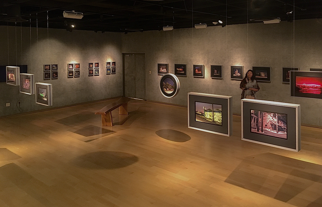 Gallery with framed artwork on walls, a person observing, wooden benches, and spotlights on ceiling.