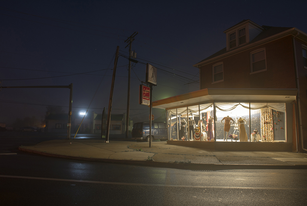 Nighttime scene of a corner building with a brightly lit storefront displaying mannequins and clothing inside.