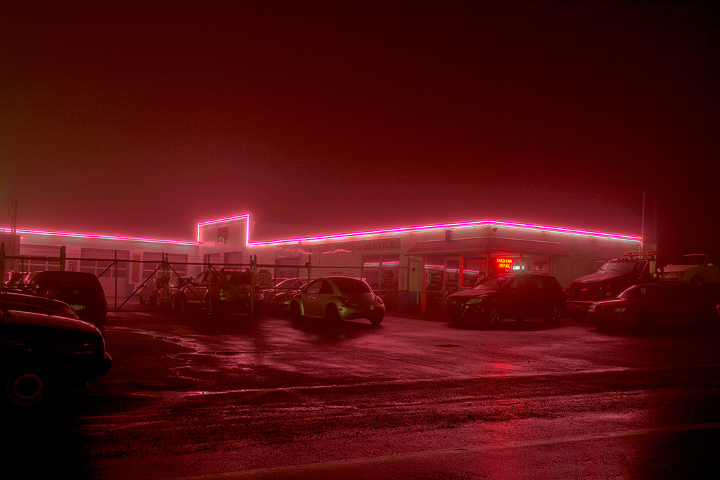 Parking lot with cars in front of a building illuminated by pink neon lights at night.