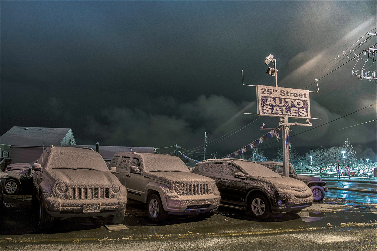 Four cars parked in a lot under dark, stormy sky with a sign for auto sales nearby.