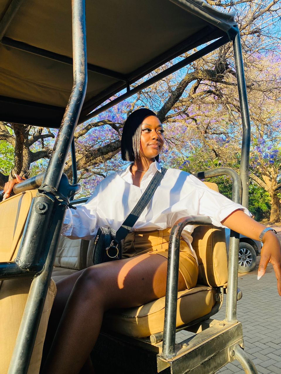 Woman sitting in a golf cart outdoors, wearing a white shirt and black tights, with trees in the background.