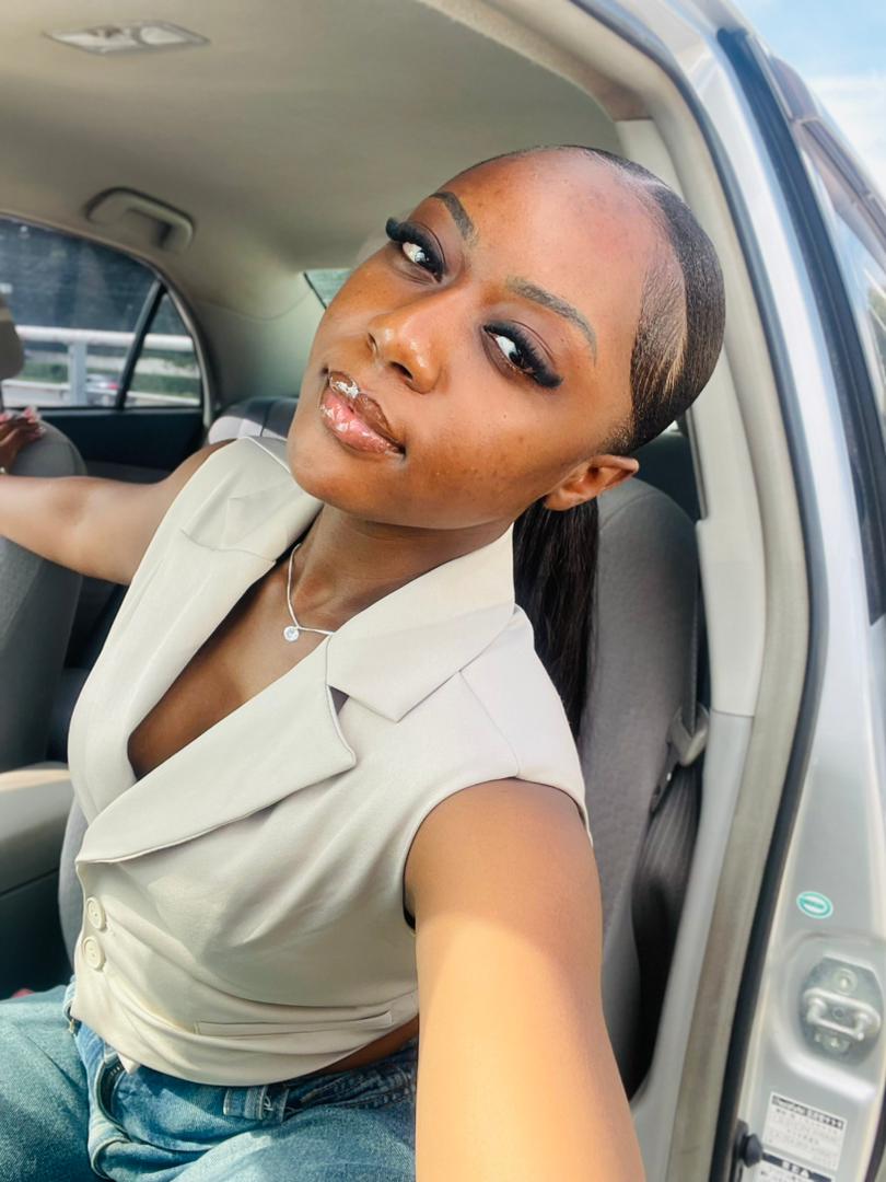 Woman with makeup and a ponytail sitting in a car, looking at the camera, wearing a light-colored top.