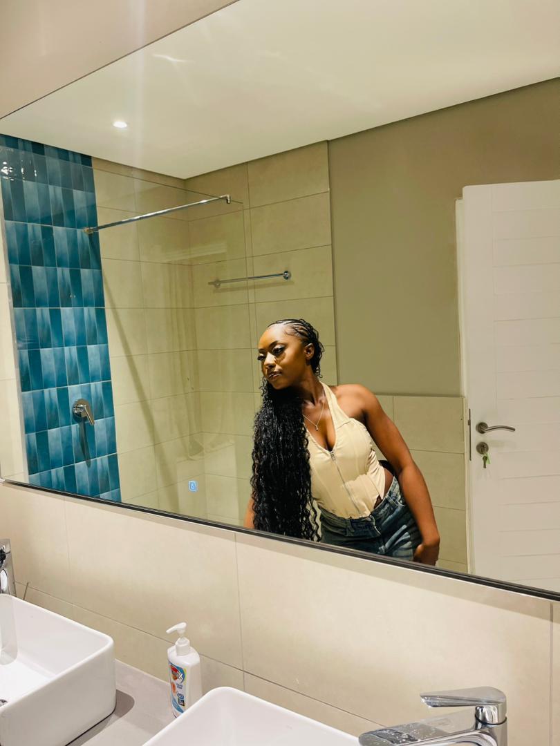 Woman with long curly hair taking a mirror selfie in a bathroom, wearing a beige top and dark pants.