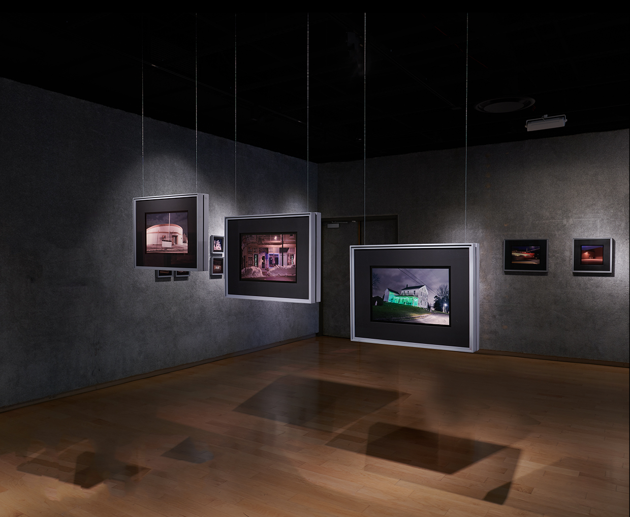 Art gallery with framed photographs hanging on dark gray walls and wooden floor, illuminated by spotlights.
