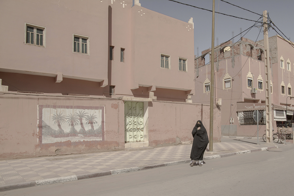 Person wearing black walking on sidewalk in front of pink buildings with windows and a mural of palm trees.