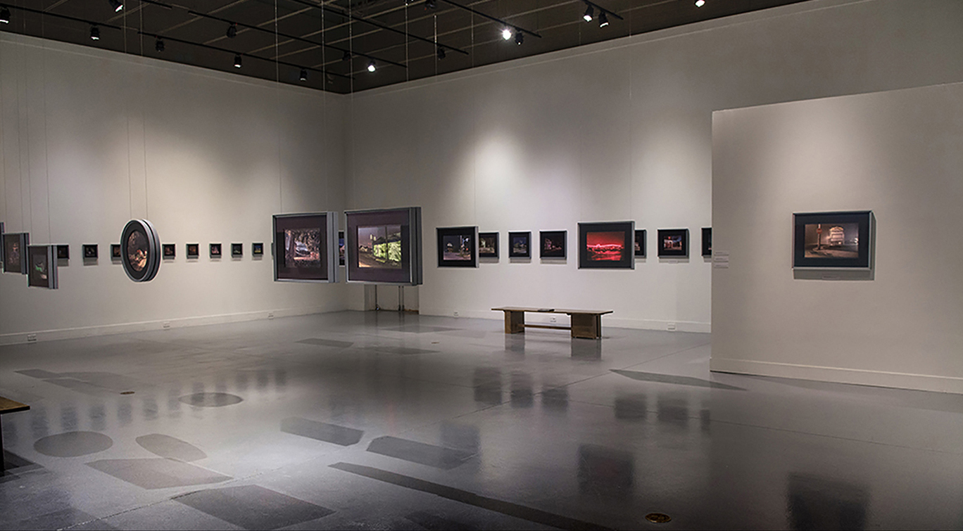 Gallery with framed artwork on white walls, benches, and a reflective floor, illuminated by ceiling lights.