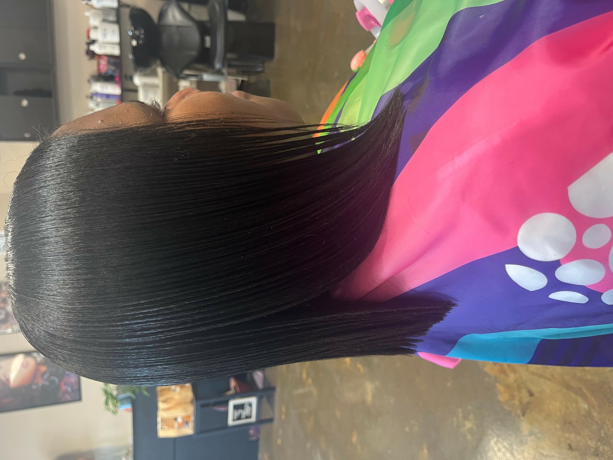 Person with long, straight, black hair in a salon chair, wearing a colorful cape with pink, green, and purple sections.