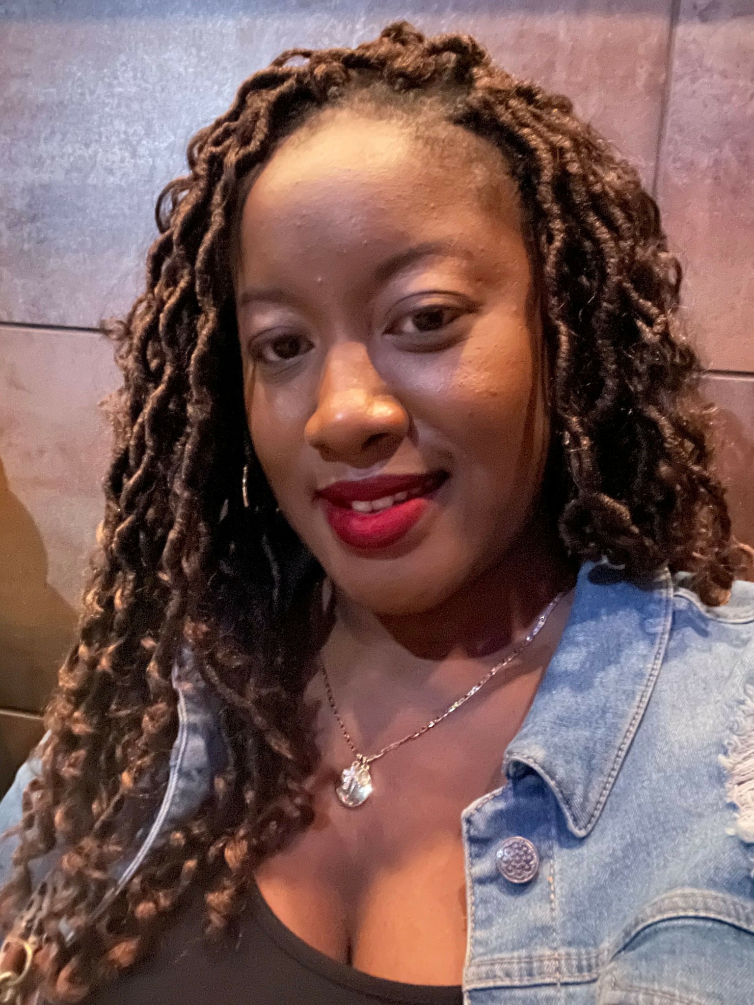 Woman with long, curly hair, wearing a denim jacket and red lipstick, smiling at the camera.