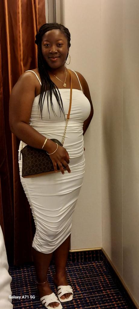 Woman in white dress standing in a narrow hallway, smiling, with a purse and jewelry.
