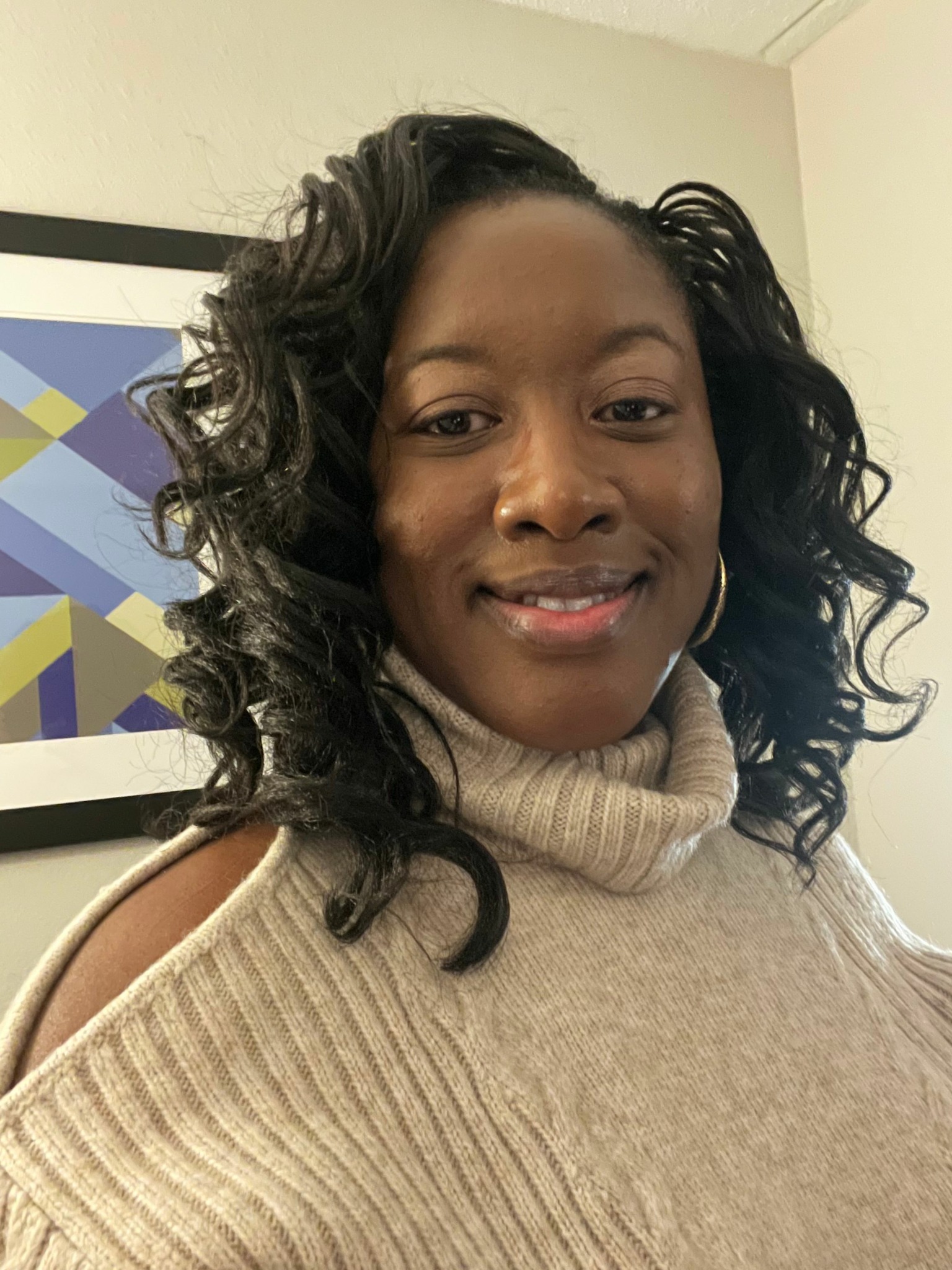 Woman with curly hair wearing a beige sweater, smiling, in front of a colorful geometric background.